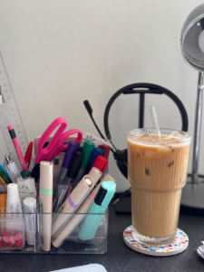 A desk setup with colorful pens, scissors, and stationery in holders beside an iced coffee in a tall, ribbed glass. A lamp and headphones sit in the background.